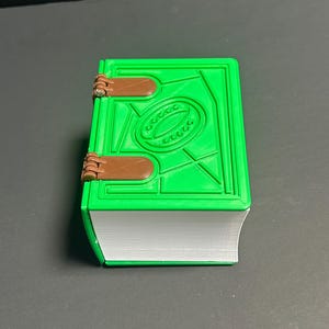 May include: A bright green, 3D-printed book with a detailed cover design and brown clasps. The book's pages are white, and the cover features an oval design. The book is a unique decorative item.