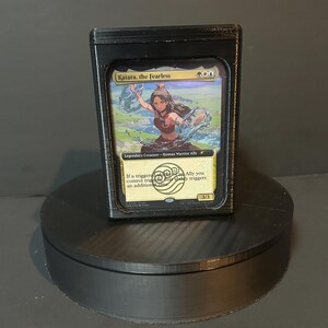 May include: A black card holder containing a Magic: The Gathering card. The card features an illustration of Katara, the Fearless, a character from Avatar: The Last Airbender. The card is set against a black background.