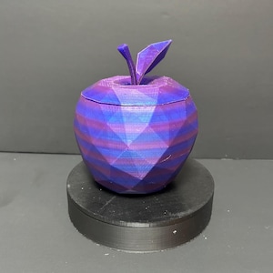 May include: A geometric apple-shaped container in shades of purple and blue, with a stem and leaf detail. The container is displayed on a black circular base. The apple design features a faceted surface.