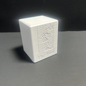 Puede incluir: Una caja blanca rectangular con un relieve detallado de un diseño de carta del tarot. La imagen presenta una figura y el sol, con el texto "The Sun" en la parte inferior. La caja está hecha de un material con líneas horizontales visibles.