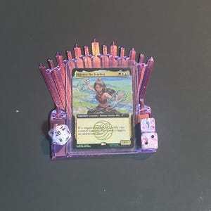 May include: A purple 3D-printed card holder, shaped like a throne, displaying a trading card of "Katara, the Fearless." Two dice are positioned on either side of the card. One die shows the numbers 8, 14, and 20. The card holder is a collectible.