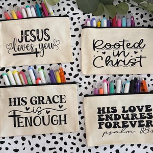 May include: Four beige canvas pencil cases with black zippers and various inspirational quotes in black script. Each case is filled with colorful pens and markers. The quotes include "Jesus loves you," "Rooted in Christ," "His grace is enough," and "His love endures forever psalm 118:1."