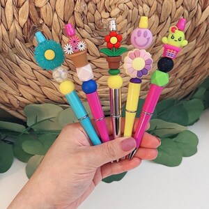 May include: A collection of colorful pens with decorative toppers. Each pen features a different design, including flowers, smiley faces, and a cactus. The pens have a variety of colors, including blue, pink, yellow, and green. The pens are held in a hand.