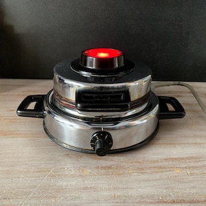 Puede incluir: Una máquina de hacer gofres cromada vintage con asas negras y un mando. La parte superior tiene una sección negra con una luz roja. El aparato está sobre una superficie de madera de color claro.