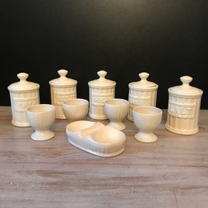 Op de afbeelding: Set van zes witte keramische kruidenpotten met deksels en zes witte keramische eierdopjes en een witte keramische eierdopjeshouder. De potten zijn gelabeld met "Tijm", "Peterselie", "Oregano", "Basilicum" en "Zout".