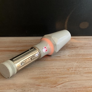 Op de afbeelding: Een vintage zaklamp met een witte en zilveren behuizing. De zaklamp heeft een cilindrisch ontwerp met een getextureerde grip en een facetkop. Een oranje gloed komt uit de kop. Een kleine rood-witte sticker is zichtbaar.