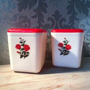 Op de afbeelding: Twee witte vierkante potten met rode deksels en een bloemenontwerp met rode rozen.