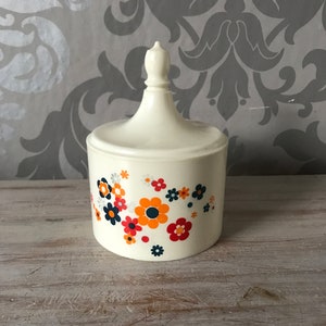 Può includere: Vaso in ceramica bianca con un motivo floreale. I fiori sono arancioni, rossi, blu e bianchi. Il vaso ha un coperchio con un pomo in cima.