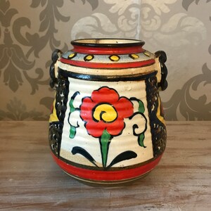 Op de afbeelding: Een witte keramische vaas met een rood, geel en groen bloemenontwerp. De vaas heeft twee handvatten en een bruine rand.