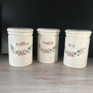 Op de afbeelding: Drie witte keramische potten met bloemmotieven en bruine letters. De potten zijn gelabeld met "koffie", "suiker" en "thee".