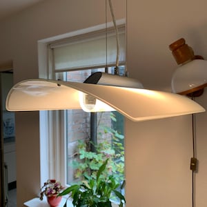 Op de afbeelding: Een witte plafondlamp met een gebogen, witte glazen kap. De lamp hangt aan een wit plafond.