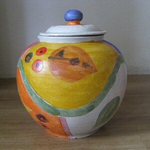 Op de afbeelding: Een keramische pot met een deksel, beschilderd met abstracte vormen en kleuren, waaronder geel, oranje, groen en rood. De pot heeft een witte achtergrond en een blauwe knop op het deksel.