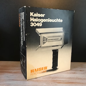 Puede incluir: Una caja negra con texto blanco que dice "Kaiser Halogenleuchte 3049". La caja muestra una luz halógena plateada con un mango negro. La parte inferior de la caja tiene el texto "KAISER FOTOTECHNIK" en blanco.
