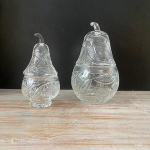 Op de afbeelding: Twee heldere, peervormige glazen containers met deksels. Elk stuk is voorzien van een gestructureerd patroon van bladeren en fruit in het persglas. Het glas is transparant en reflecterend, met een duidelijke steel aan de bovenkant van elk deksel. De set bevat een kleinere en een grotere peervormige pot.
