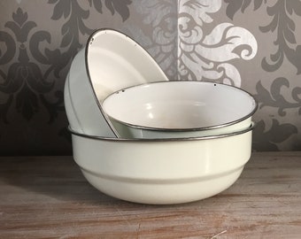 May include: Three white enamel bowls with black rims are stacked on top of each other. The bowls are on a wooden surface.