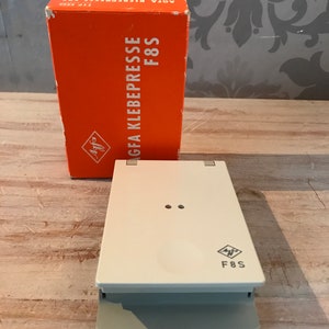 Op de afbeelding: Een oranje doos met de tekst "AGFA KLEBEPRESSE F8S" en het Agfa-logo. De doos staat op een houten oppervlak. Een witte en grijze Agfa F8S lijm pers staat op het houten oppervlak.