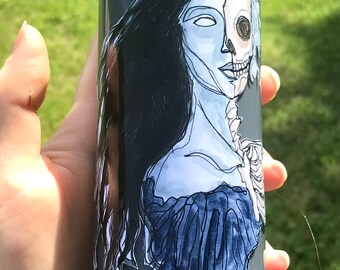 May include: A gray glass candle with a white sticker of a woman with a skull face. The woman has long black hair and is wearing a blue dress.
