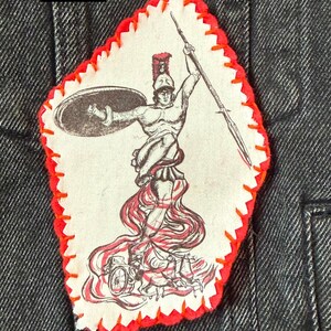 May include: A patch featuring a detailed illustration of Ares, the Greek god of war, holding a spear and shield. The patch has a red stitched border and the word "Ares" in a black box. The patch is attached to a dark denim jacket.