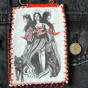 May include: A rectangular patch with a stitched red border and a safety pin. The patch features a black and white illustration of three figures with wolves, and the word "Hekate" in a black rectangle.