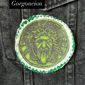 May include: A round patch featuring a green and black Gorgoneion design, with a white and green scalloped border. The patch is affixed to a dark denim jacket. The word "Gorgoneion" is displayed in a black rectangle.