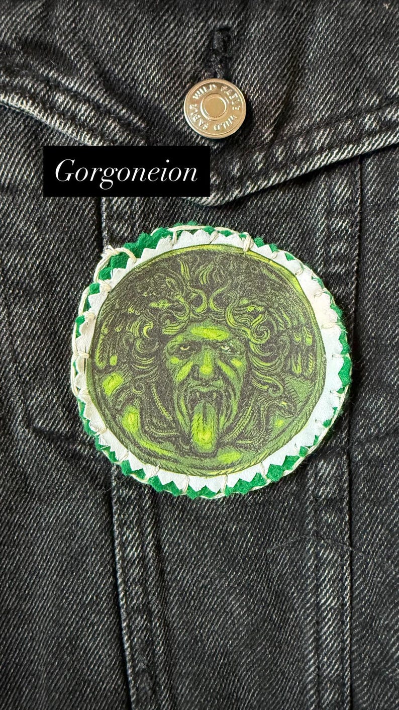 May include: A round patch featuring a green and black Gorgoneion design, with a white and green scalloped border. The patch is affixed to a dark denim jacket. The word "Gorgoneion" is displayed in a black rectangle.
