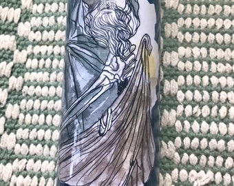 May include: A dark blue candle with a detailed illustration of a robed figure with a long beard. The artwork is in shades of gray and white, with the figure holding a staff or similar object. The candle has a glossy finish.