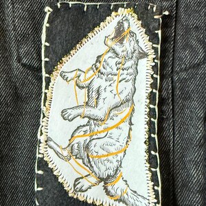 May include: A black denim jacket with a patch featuring a howling wolf illustration. The patch has a white background with a detailed drawing of a wolf bound by gold-colored ropes. The word "Fenrir" is printed in white on a black rectangle.