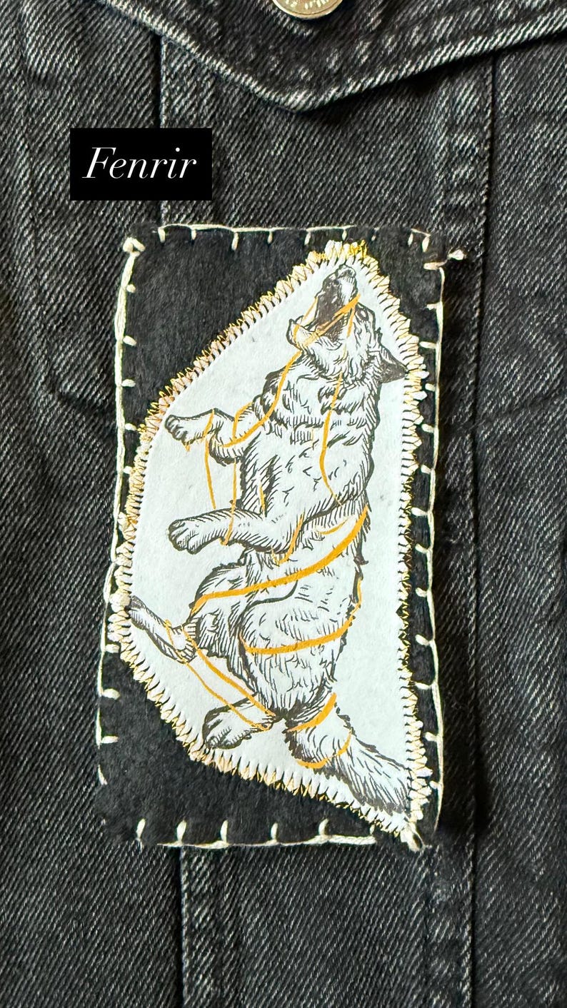 May include: A black denim jacket with a patch featuring a howling wolf illustration. The patch has a white background with a detailed drawing of a wolf bound by gold-colored ropes. The word "Fenrir" is printed in white on a black rectangle.