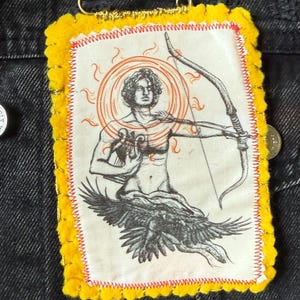 May include: A fabric patch with the word "Apollo" in a black rectangle. The patch features an illustration of a man with a bow and arrow, surrounded by a sunburst design, and an eagle. The patch has a yellow border with red stitching.