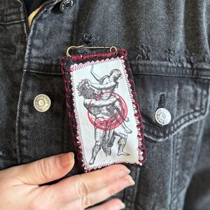May include: A rectangular fabric patch with a red and black stitched border, featuring a black and white illustration of two figures in a dynamic pose, with a red circular accent. The patch is attached to a black denim jacket with a safety pin.