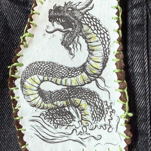 May include: A fabric patch featuring a detailed black and yellow dragon illustration. The patch has a brown border with green stitching and the text "Jormungandr" in a black rectangle.