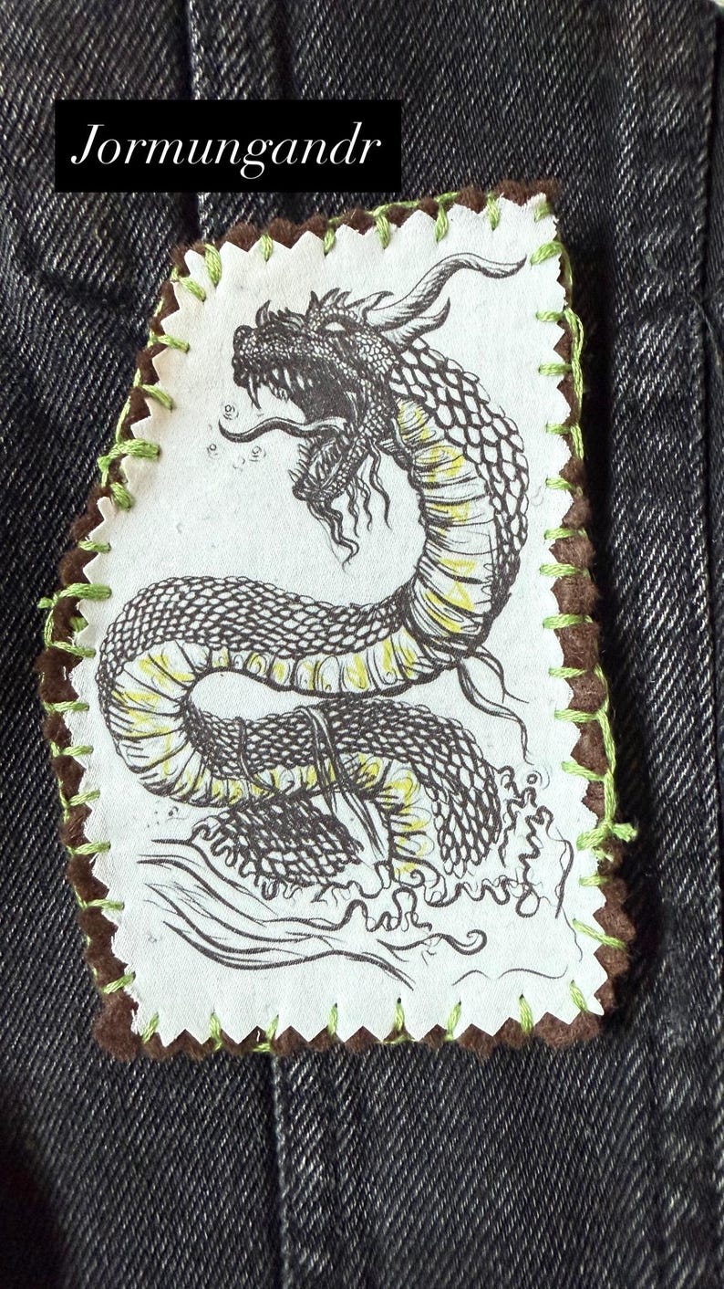 May include: A fabric patch featuring a detailed black and yellow dragon illustration. The patch has a brown border with green stitching and the text "Jormungandr" in a black rectangle.