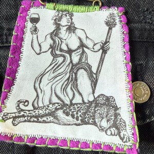 May include: A fabric patch featuring a black and white illustration of Dionysus holding a wine glass and staff, with a leopard. The patch has a purple border with green stitching and the word "Dionysus".
