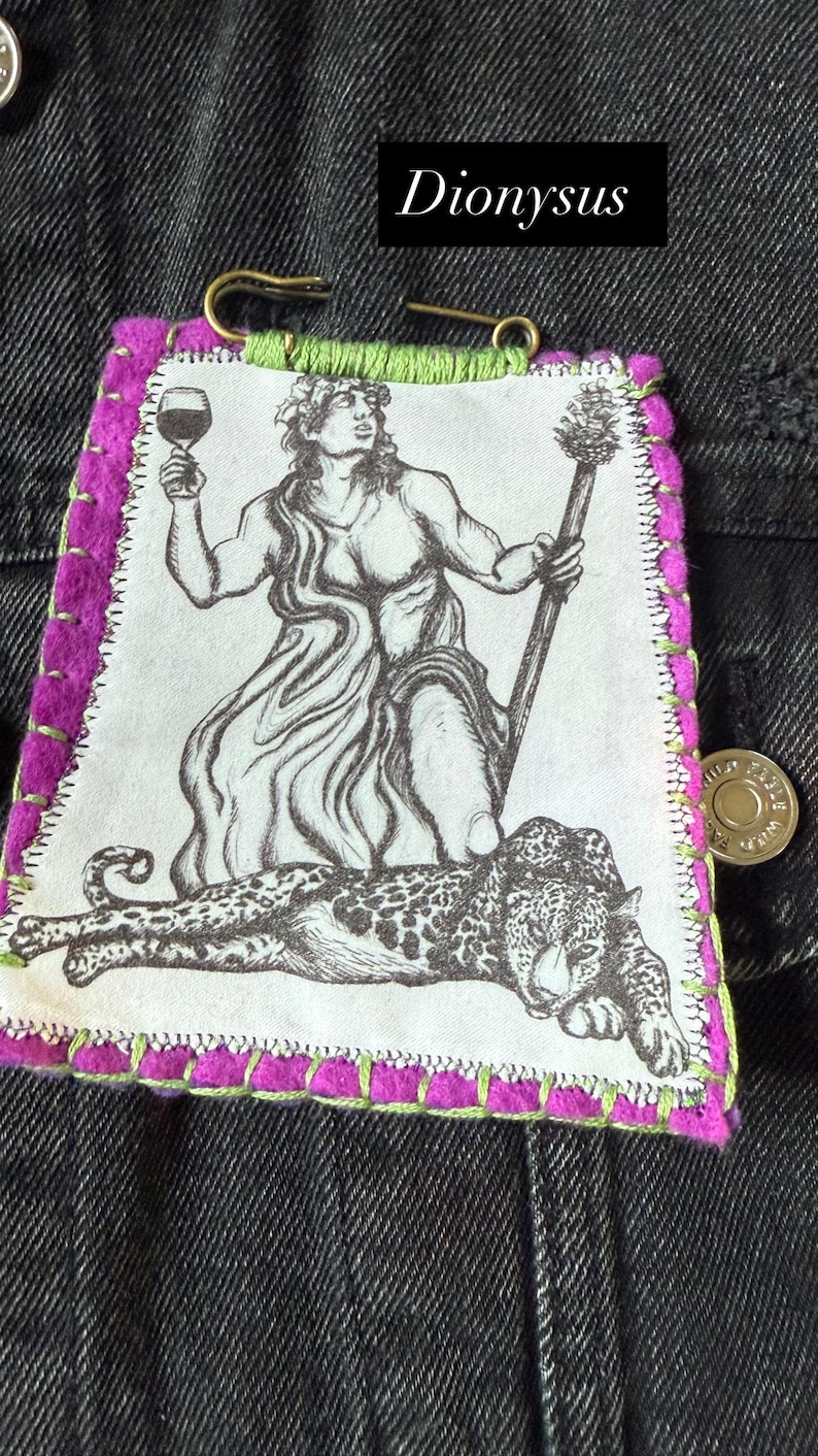 May include: A fabric patch featuring a black and white illustration of Dionysus holding a wine glass and staff, with a leopard. The patch has a purple border with green stitching and the word "Dionysus".