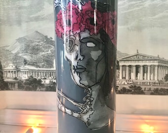 May include: A gray candle with a black and white illustration of a woman's face with a floral crown. The woman's face is partially obscured by a skull.