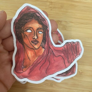 Op de afbeelding: Een sticker met een aquarel-illustratie van een vrouwenhoofd met een rode kap. De ogen van de vrouw zijn wit en haar gezicht is lichtbruin.