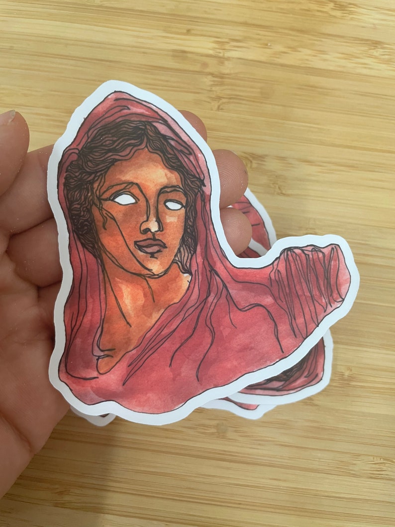 Op de afbeelding: Een sticker met een aquarel-illustratie van een vrouwenhoofd met een rode kap. De ogen van de vrouw zijn wit en haar gezicht is lichtbruin.