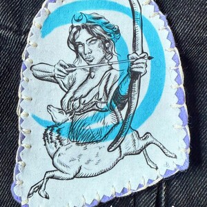 May include: A fabric patch featuring a black and white illustration of Artemis, the Greek goddess of the hunt, with a bow and arrow, riding a deer. The patch has a light blue and purple border and the name "Artemis" is printed at the bottom.