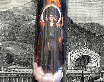 May include: A tall, cylindrical candle with a clear top and a decorative wrap featuring a celestial design. The wrap depicts a woman with three faces, holding a crescent moon, with a fiery element above. The design includes stars and a cloud base.