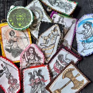 Könnte beinhalten: Eine Sammlung von Stoffaufnähern mit Illustrationen mythologischer Figuren, darunter ein Wolf, ein Bogenschütze und Medusa. Jeder Aufnäher hat einen einzigartigen farbigen Rand und ist auf einem dunklen Hintergrund aufgenäht.
