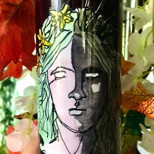 Könnte beinhalten: Eine hohe, dunkelviolette Kerze mit einer gemalten Illustration eines Frauengesichts. Das Gesicht ist geteilt, eine Seite in hellen Farben, die andere in dunklen Farbtönen. Die Frau ist mit Blumen und einer Krone geschmückt.