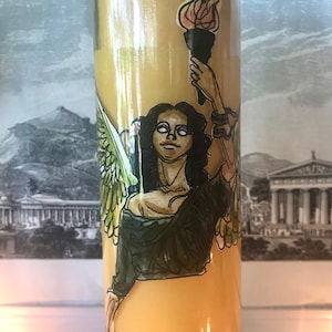 May include: A yellow candle with a sticker of a woman with wings holding a torch. The woman has long dark hair and is wearing a green dress. The background of the sticker is a black and white image of a building.
