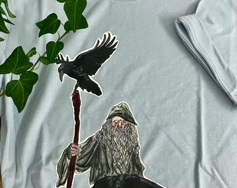 May include: Light blue t-shirt featuring a wizard with a long white beard, holding a staff, and two black ravens. The text "MORNINGSTAR WITCHERY" is printed above the wizard. The shirt is made of soft material.