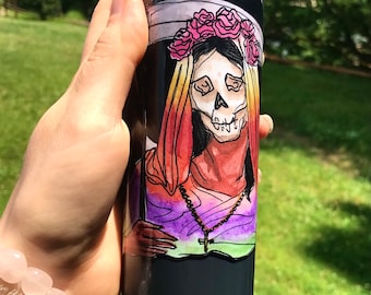 Può includere: Una candela nera con un'illustrazione ad acquerello di una figura con la faccia da teschio che indossa una corona floreale rosa e una collana con una croce. La figura è raffigurata con abiti colorati su sfondo nero.