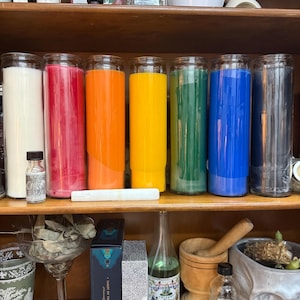 May include: A shelf displays a collection of colorful candles in glass jars, ranging from white to black. Additional items include small bottles, a mortar and pestle, and a jar labeled "MINT". The arrangement suggests a focus on spiritual or decorative items.