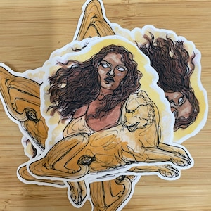 May include: A sticker featuring a woman with long brown hair and a white halo, holding a golden lion. The woman has a light brown skin tone and is wearing a white top. The sticker is on a wooden background.