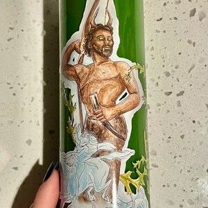 Puede incluir: Vela de vidrio verde con una pegatina que representa una figura musculosa con barba, sosteniendo un cuchillo y un vaso. La pegatina tiene un fondo blanco con detalles en azul y verde. La vela es cilíndrica.