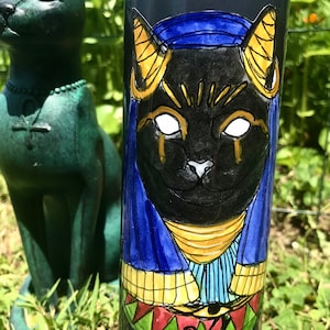 Op de afbeelding: Een zwarte kaars met een handgeschilderd beeld van een zwarte kat met gouden en blauwe accenten. De kat draagt ​​een blauwe en gouden hoofdtooi en heeft een gouden oogsymbool op zijn borst.