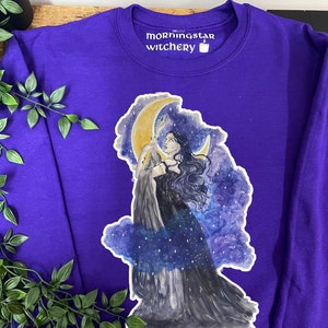 May include: A purple sweatshirt featuring a watercolor illustration of a woman with long dark hair, holding a crescent moon. The design includes a starry night sky and clouds. The text "morningstar witchery" is printed on the collar.