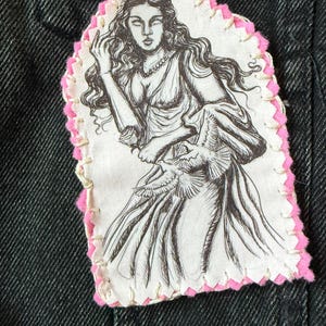 May include: A black and white illustration of Aphrodite, the Greek goddess of love, on a white patch with a pink border. The patch is sewn onto a dark denim jacket. The word "Aphrodite" is written in a black rectangle at the bottom.
